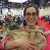 Sara Varon stands with a smile at a convention, holding a bag with a mammal with long ears inside of it.