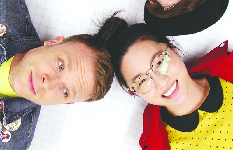 Photo of a white man in a denim jacket and an Asian woman with glasses, a yellow dress and red jacket with their heads pressed together.