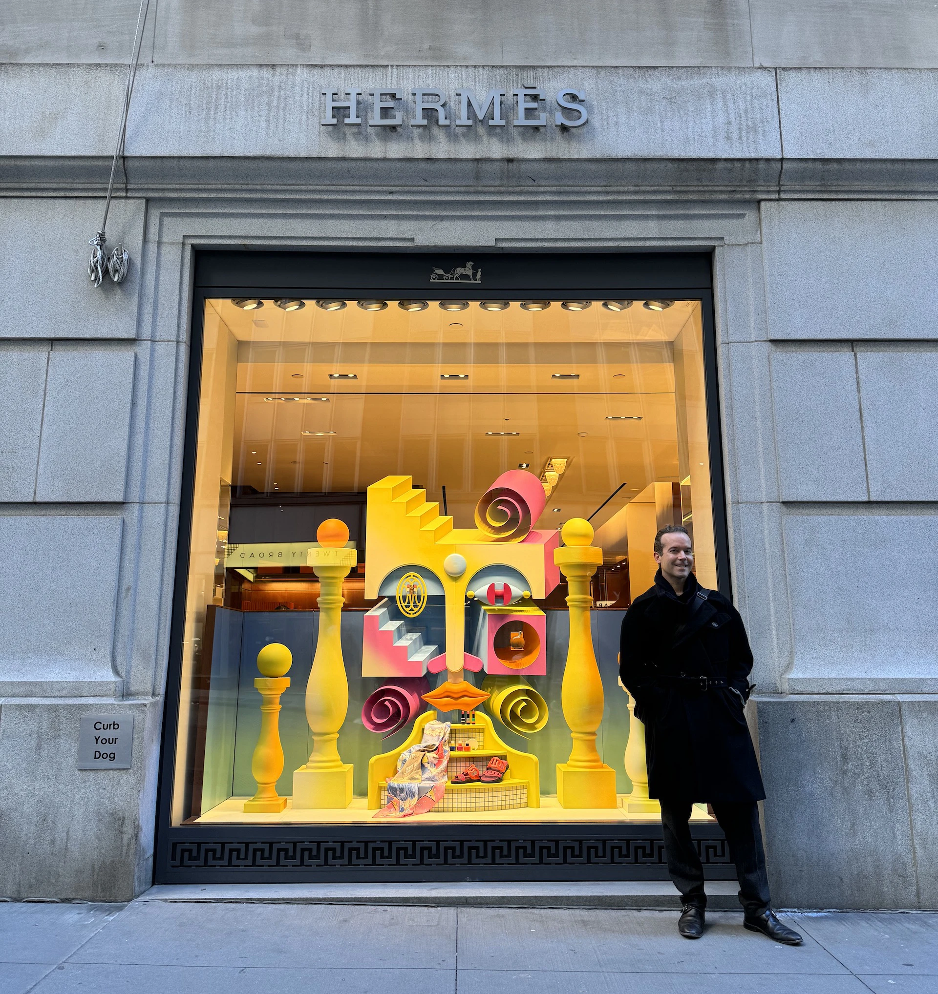 Panayiotis Terzis stands outside a building in a black coat and pants. The logo Hermes is labelled on top of the window. And in the window, a colorful window display featuring 3D geometric shapes and blocks, all artfully arranged to form a face.